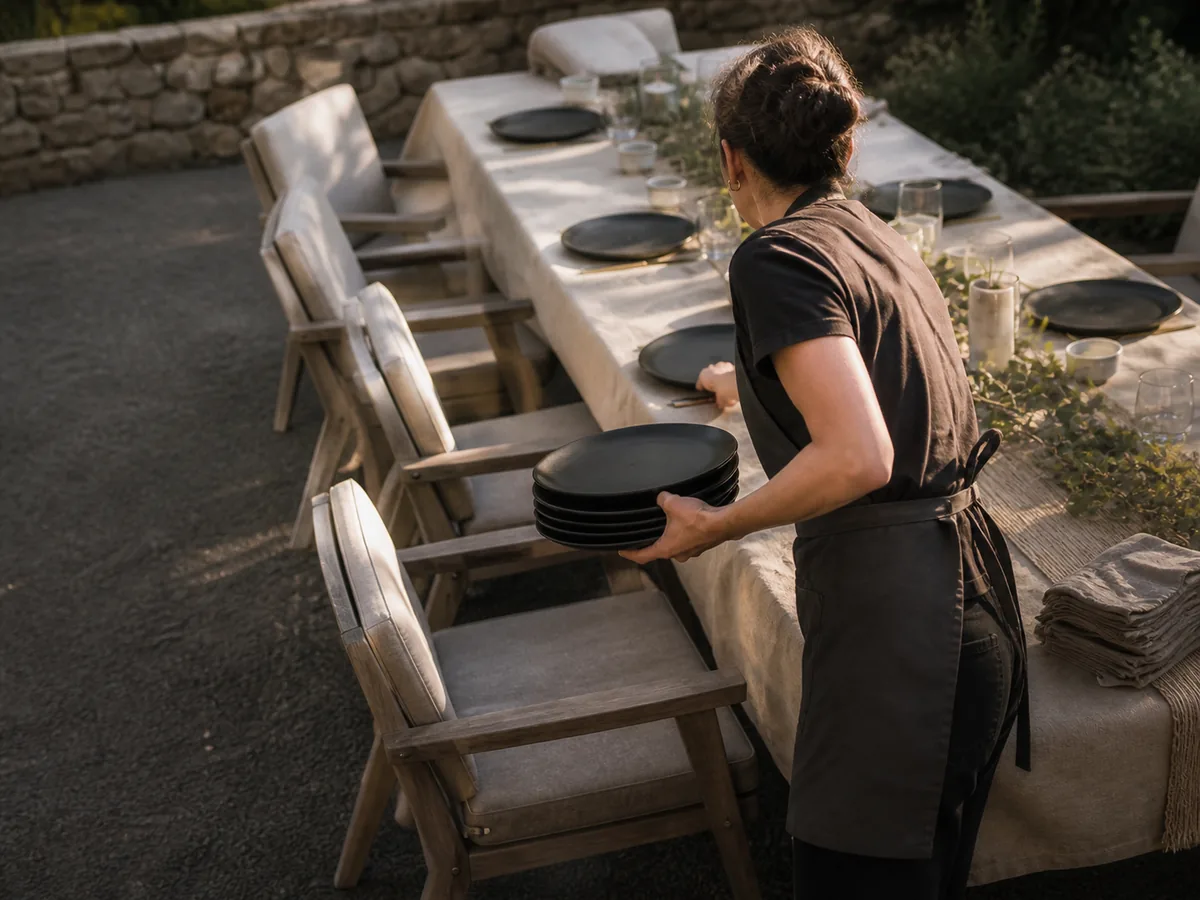 Someone in an apron laying black plates along a long linen-covered outdoor table