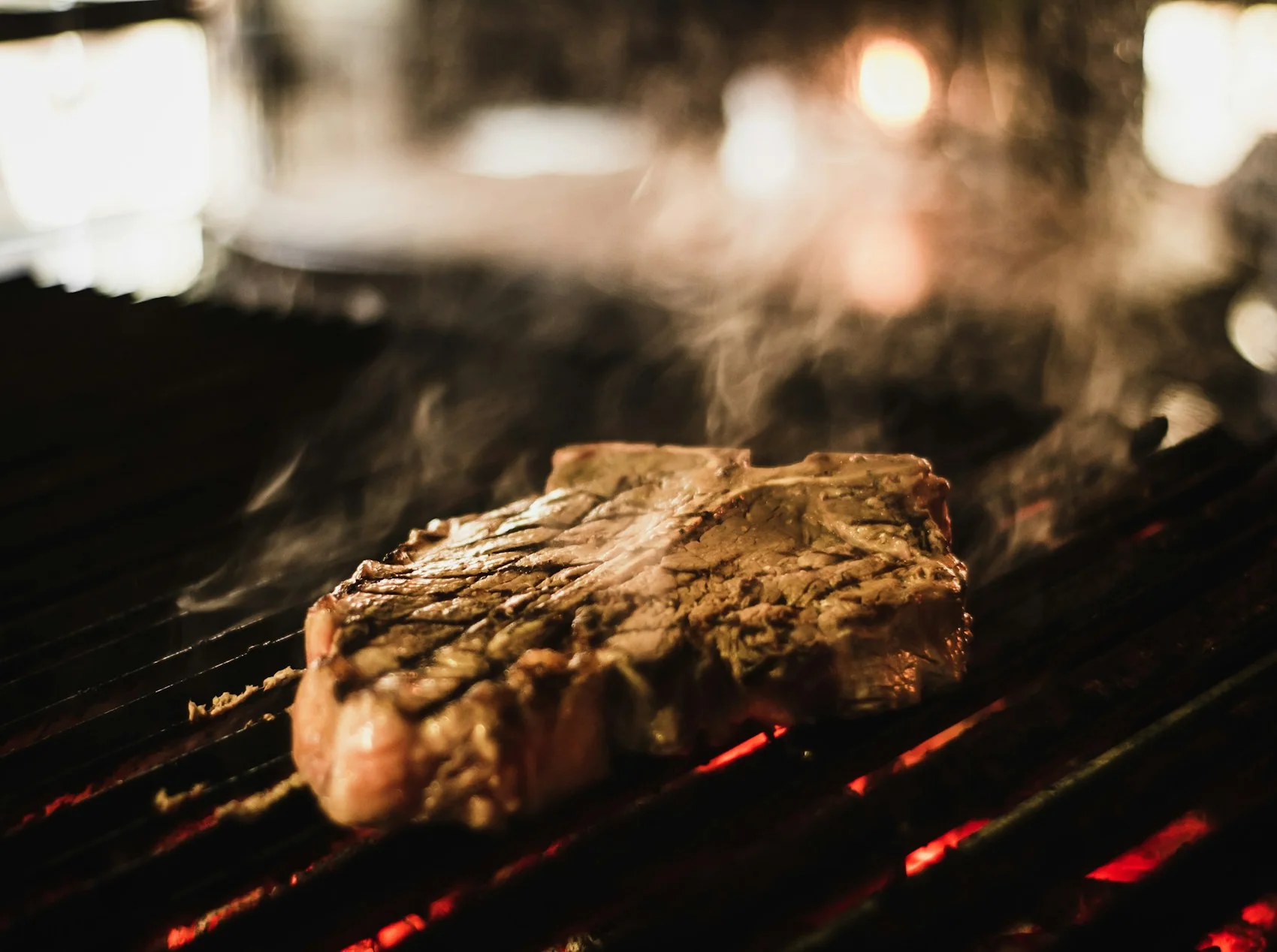 A thick steak searing over glowing coals, smoke rising off the grill