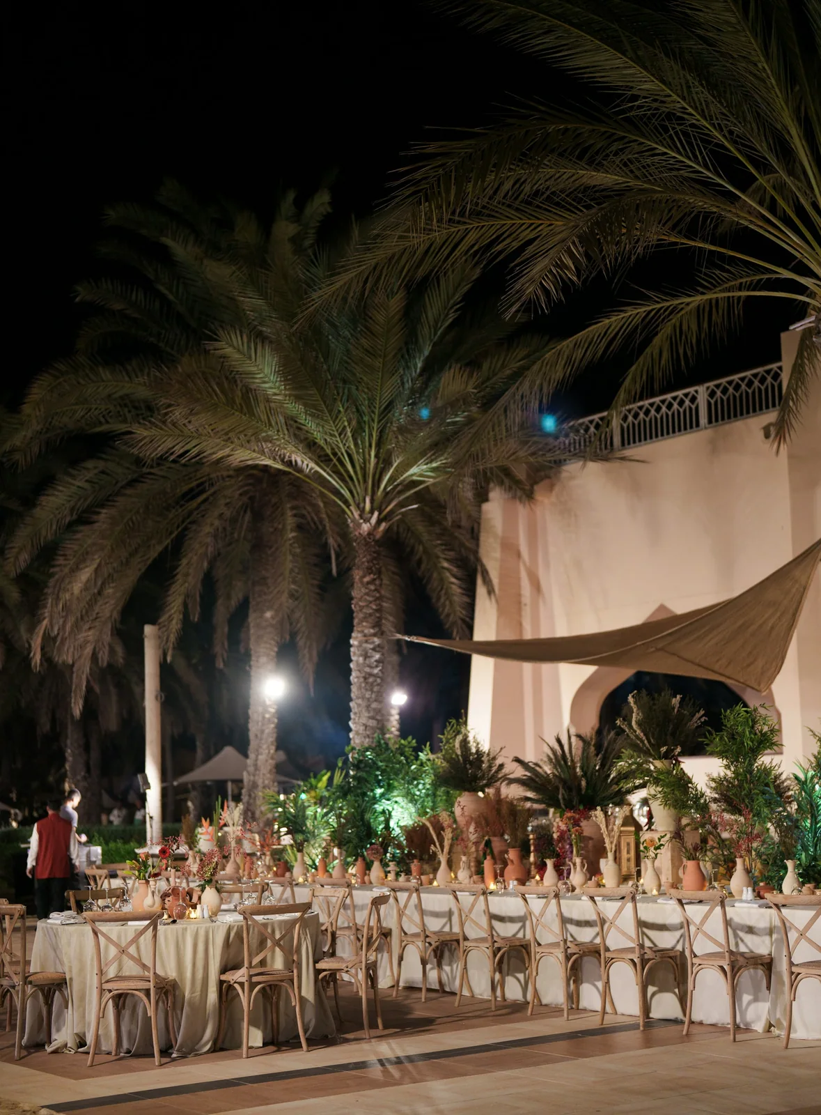 A long table set beneath palms for an evening reception, lit by lanterns