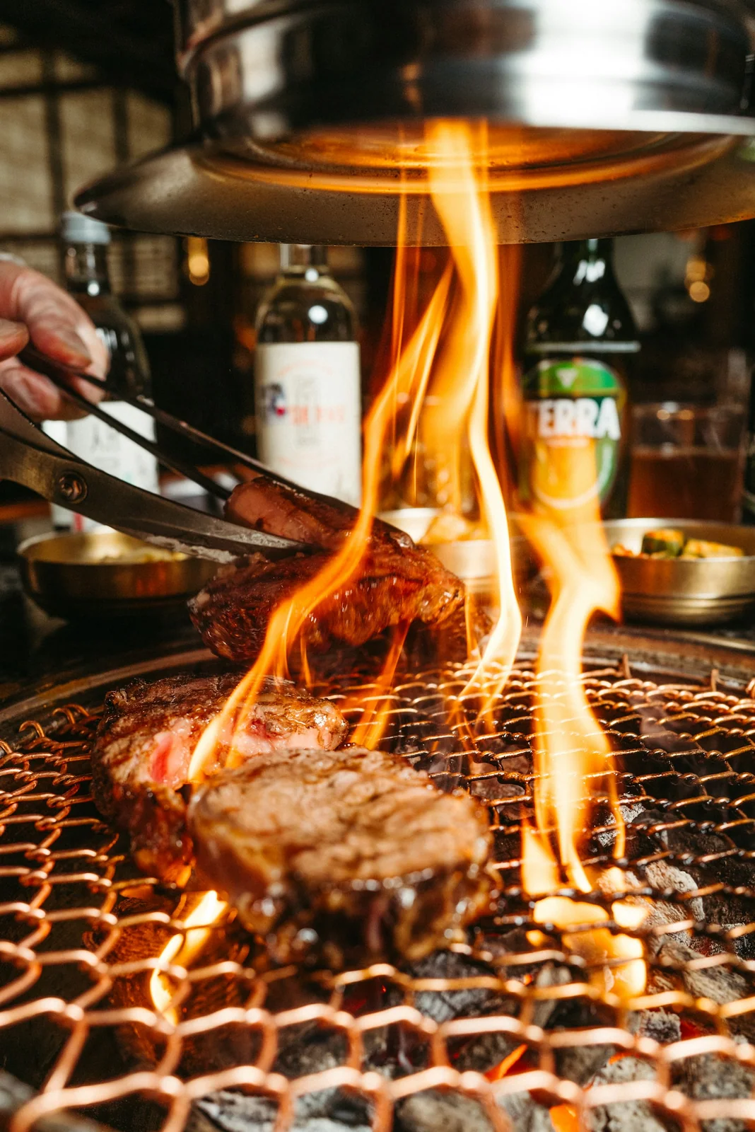 Cuts of steak flaming on a round charcoal grill as the chef turns them with tongs